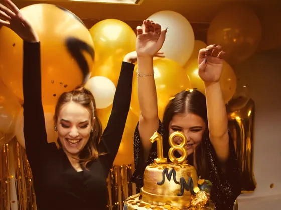 Două prietene care sărbătoresc 18 ani lângă un tort auriu cu baloane